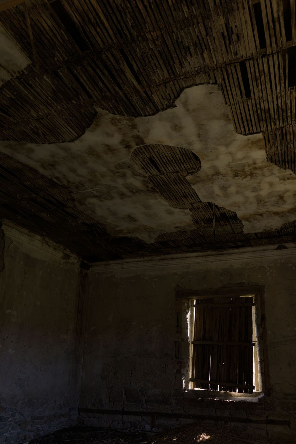 Abandoned room with deteriorating ceiling revealing wooden lath strips, peeling plaster, and a broken window letting in dim light.