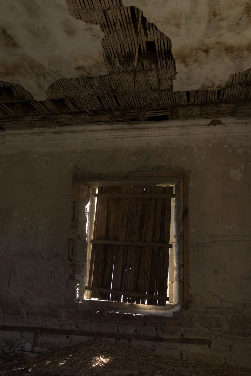 Abandoned building interior with deteriorating ceiling and boarded window letting in light through broken wooden slats.