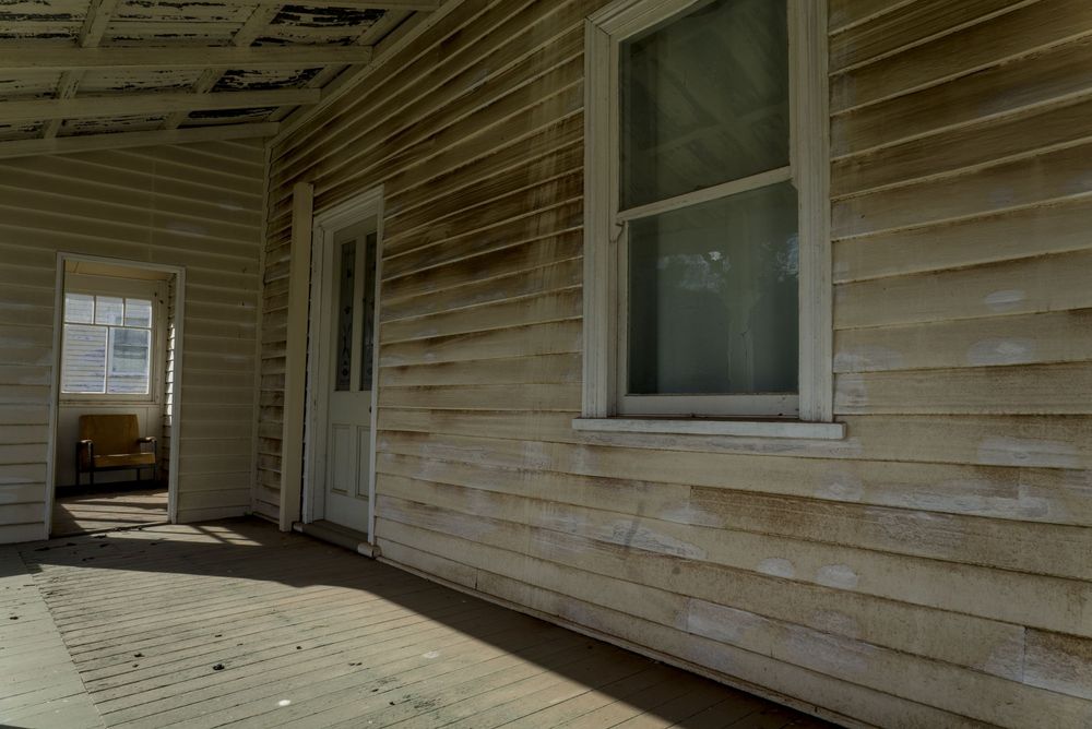 Weathered white wood siding on an old house with a covered porch, wooden deck, and white door visible.