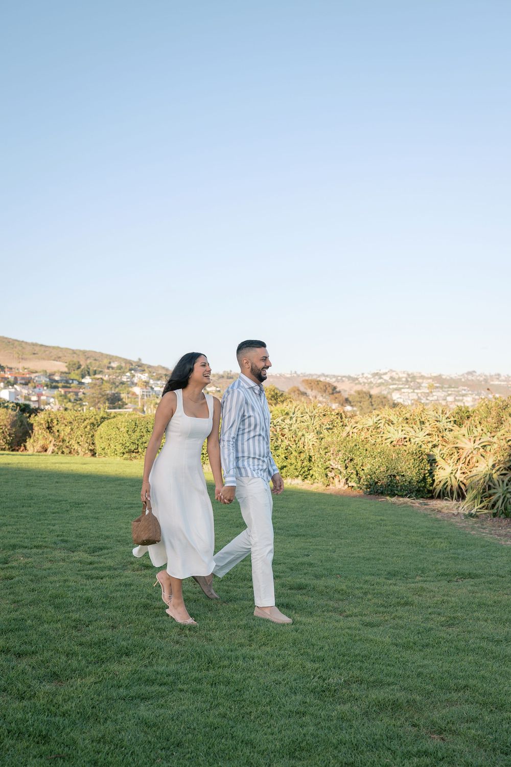 Couple holding hands walking on green grass with scenic hillside backdrop on a clear sunny day.