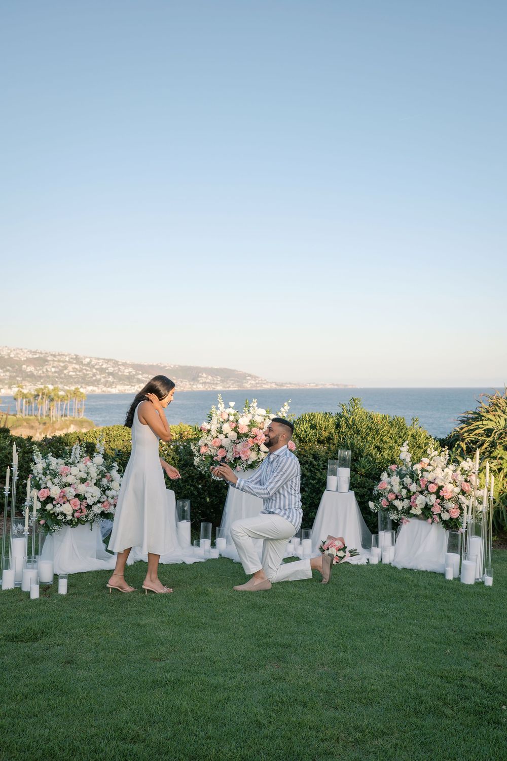 Man proposes to woman on one knee surrounded by pink floral arrangements and candles overlooking the ocean at sunset.
