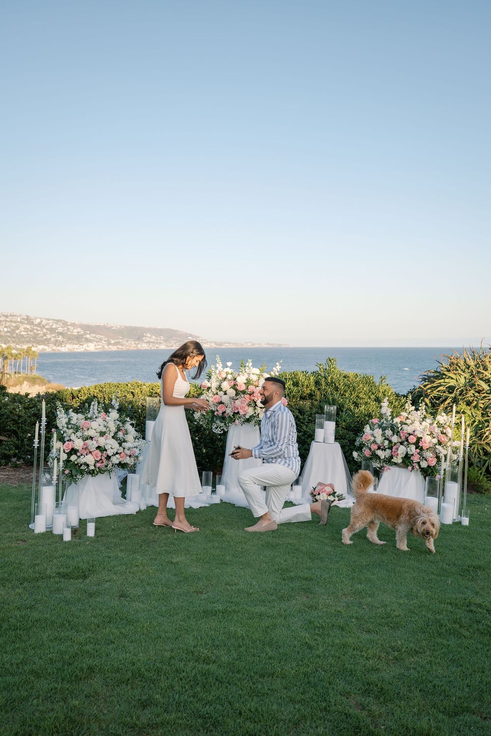 Man proposes to woman on one knee outdoors with floral arrangements, candles, ocean view, and a golden dog nearby.