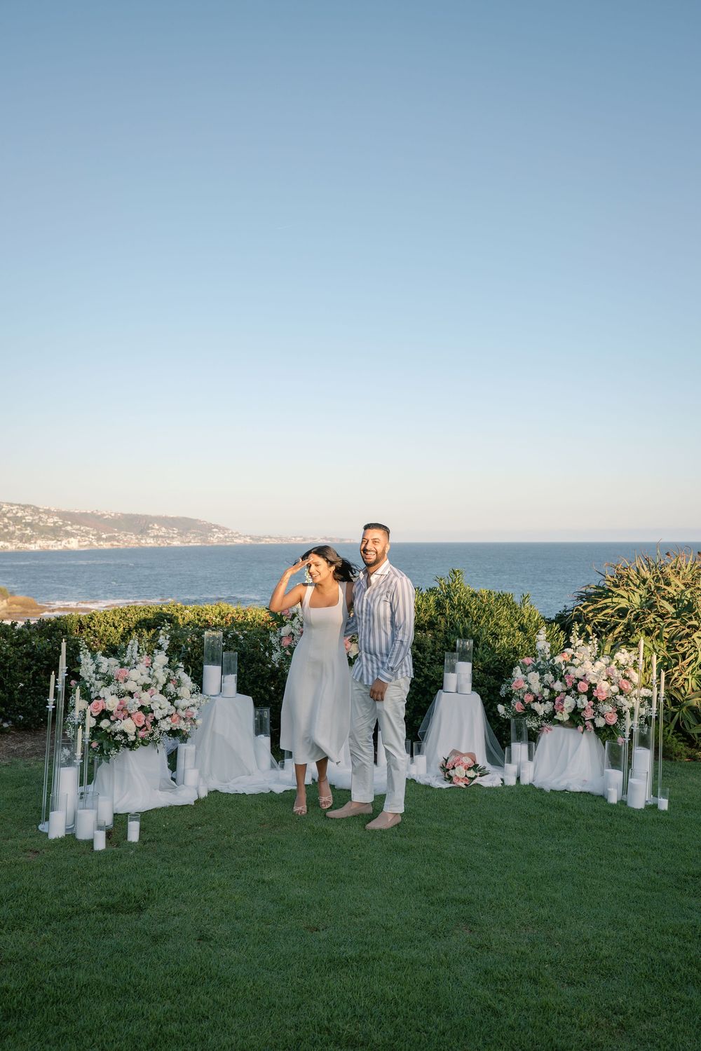 Couple standing among white candles and pink floral arrangements at a coastal outdoor proposal setup at sunset.