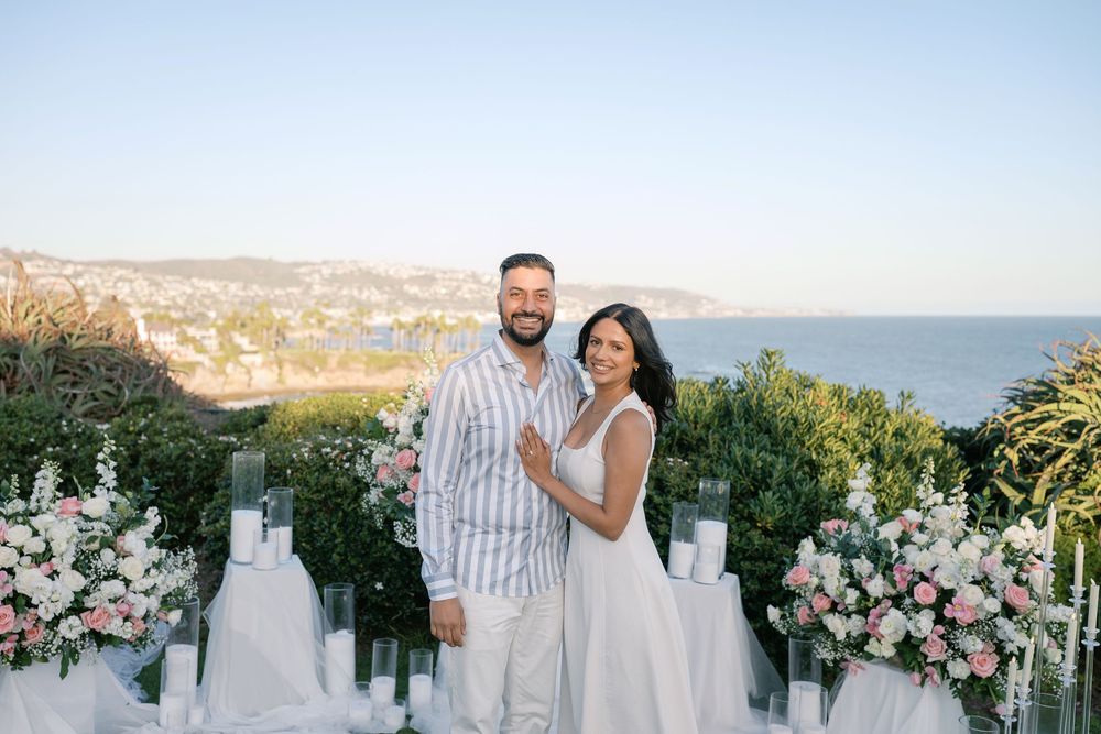Couple smiling together at an outdoor engagement setup with floral arrangements and ocean view backdrop.