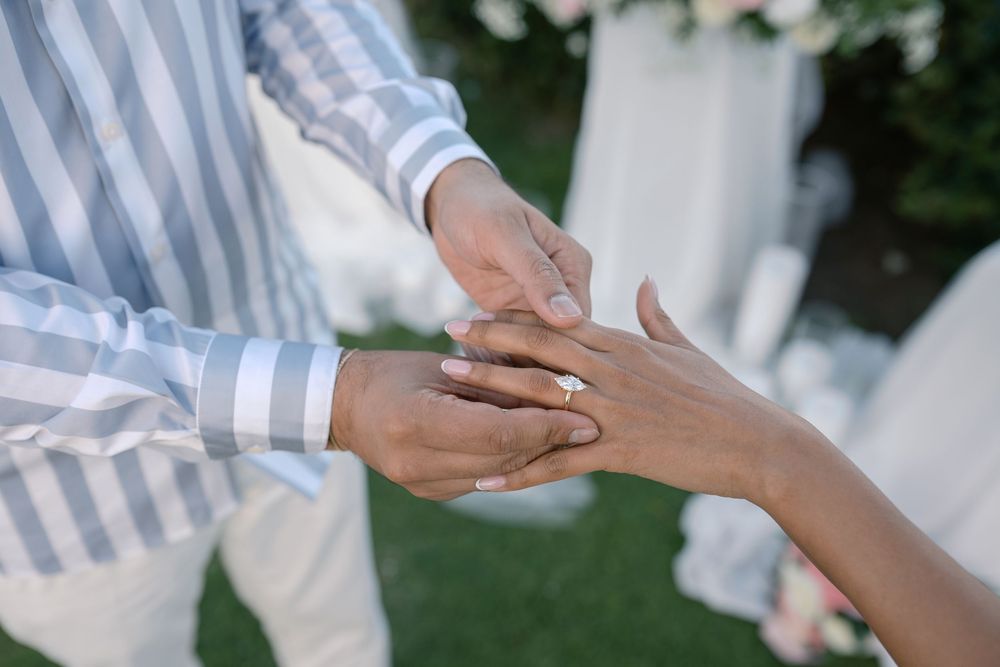 A groom places a diamond engagement ring on his bride's finger during an outdoor wedding ceremony.