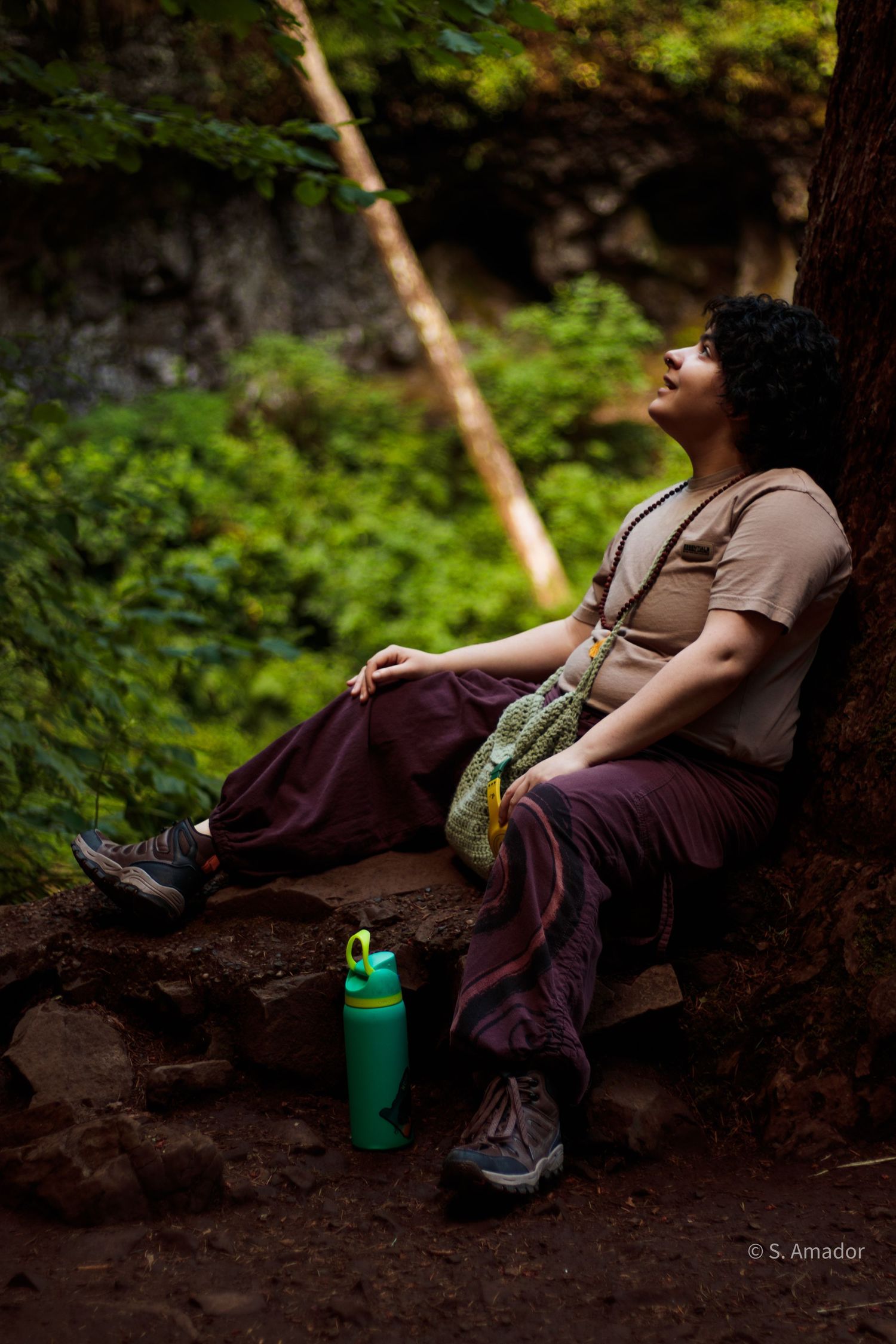 A hiker rests against a tree in a lush forest, wearing maroon pants, with a green water bottle nearby.