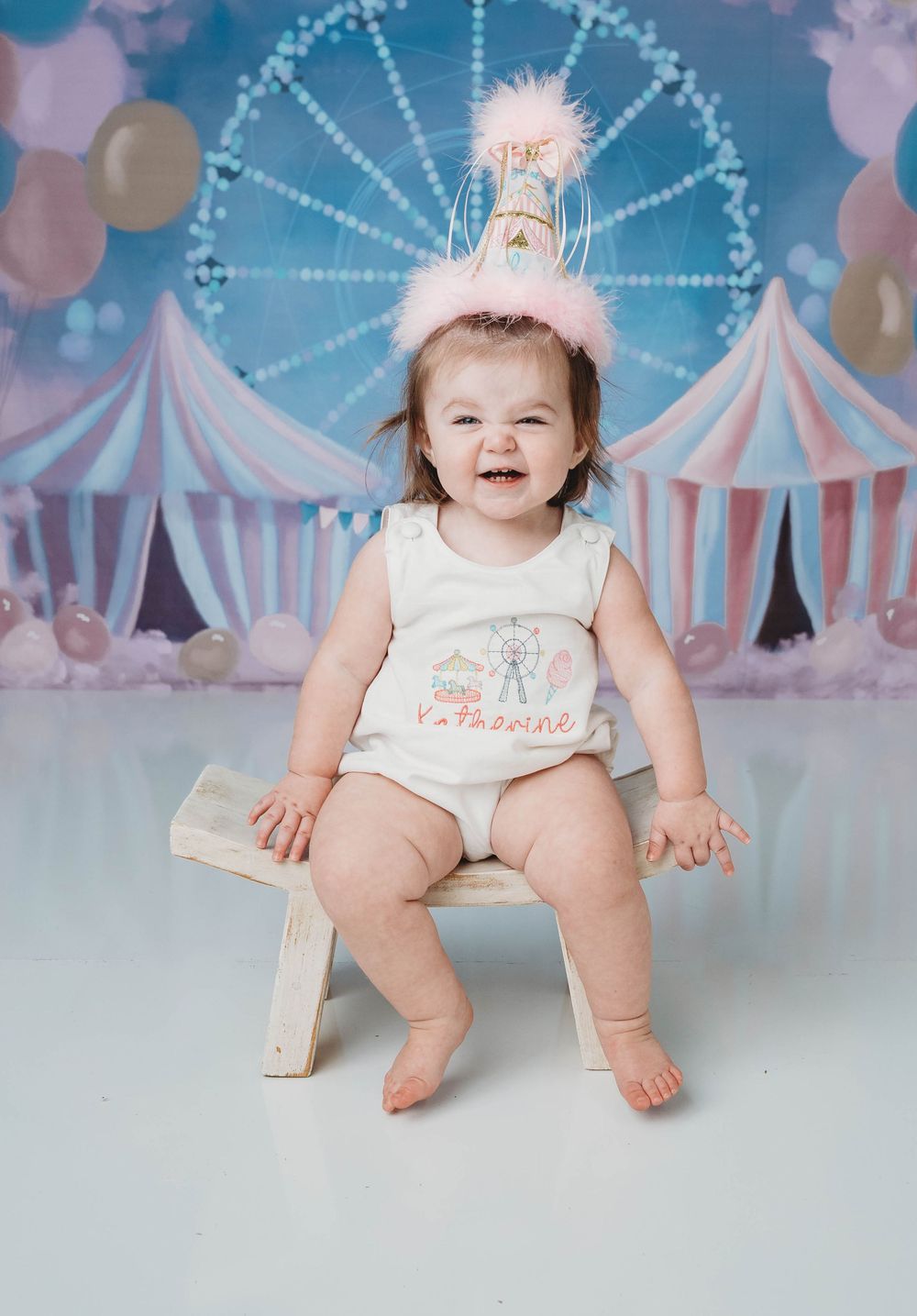 Baby girl in birthday hat sits on bench before circus backdrop for her first birthday cake smash photo.