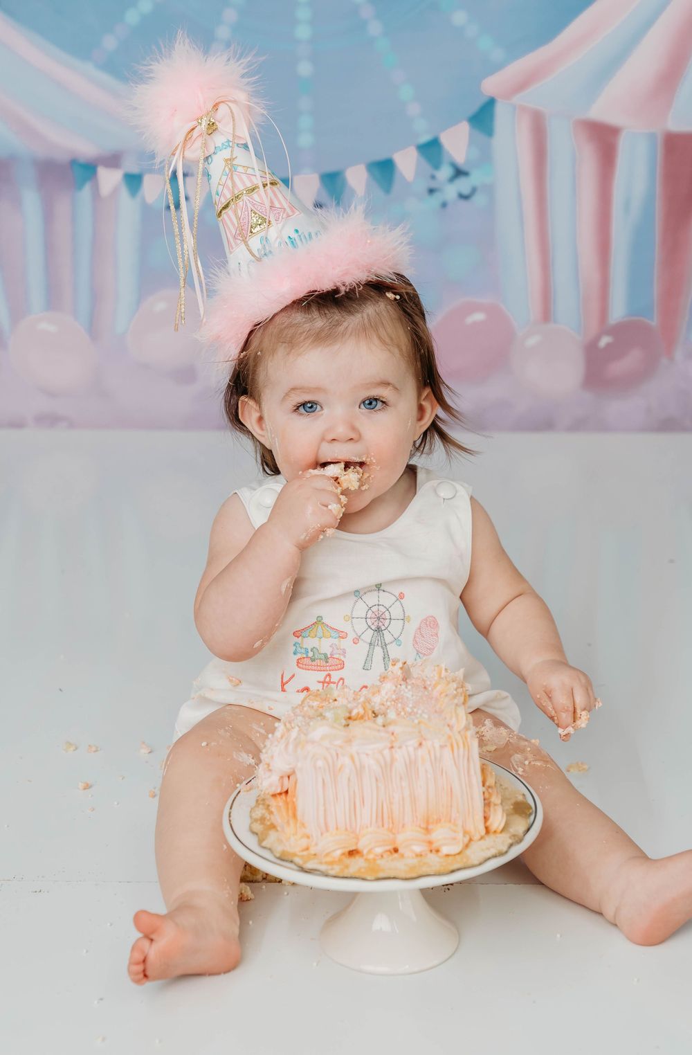 Baby girl in pink party hat smashes first birthday cake at a pastel circus-themed cake smash photo session.