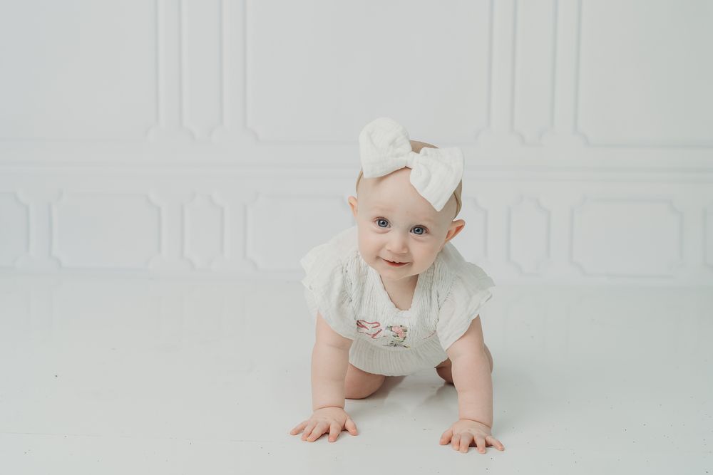 Adorable baby girl in white outfit and bow headband crawling on white floor in bright studio setting.