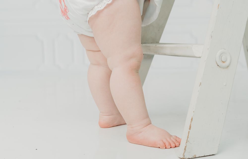 Close-up of chubby baby legs standing barefoot next to a white wooden chair on a white background.