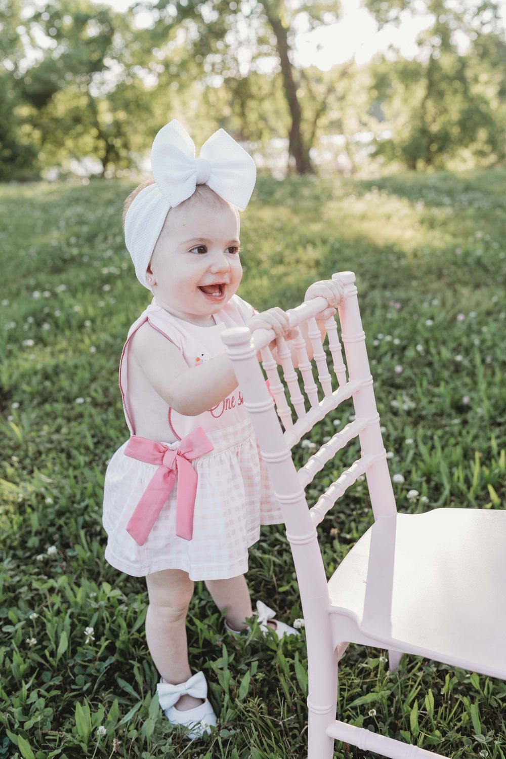 Adorable baby girl in pink dress and white bow headband standing by a pink chair on green grass outdoors.