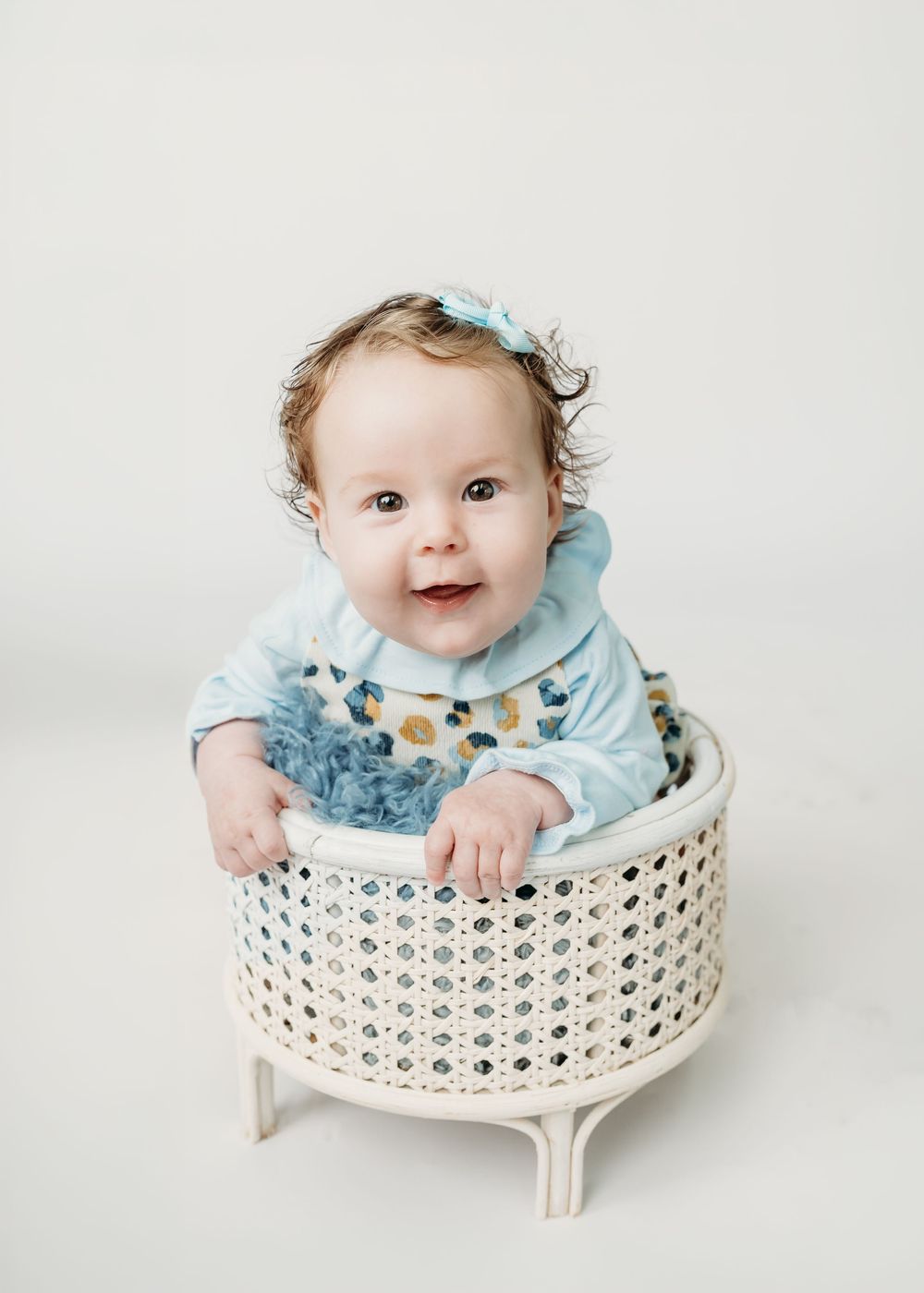 Smiling baby in blue outfit sitting in white wicker basket with blue bow headband on white background.