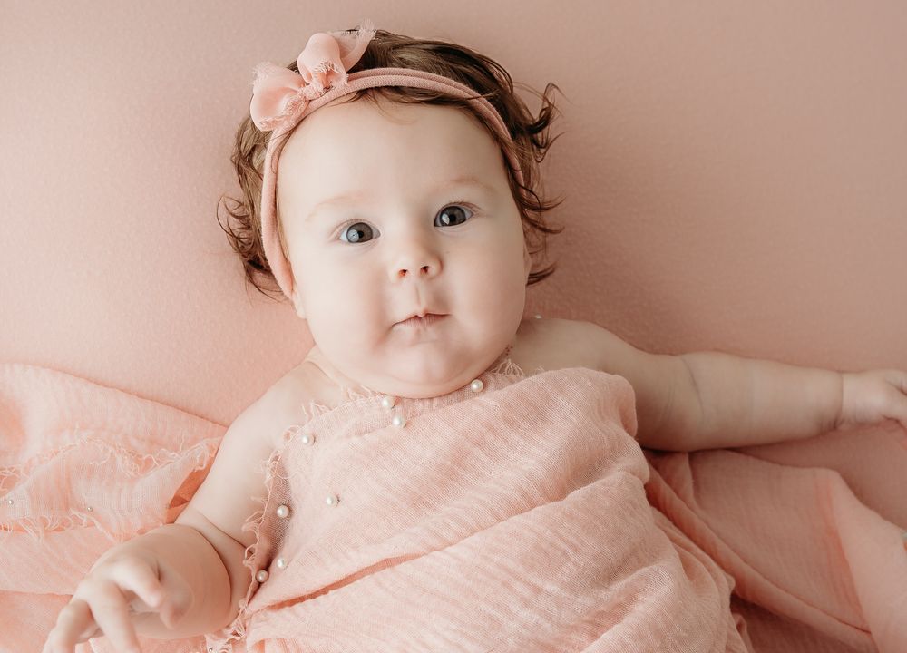 Adorable baby girl in a pink dress and bow headband lying on a soft pink blanket during a newborn photo session.