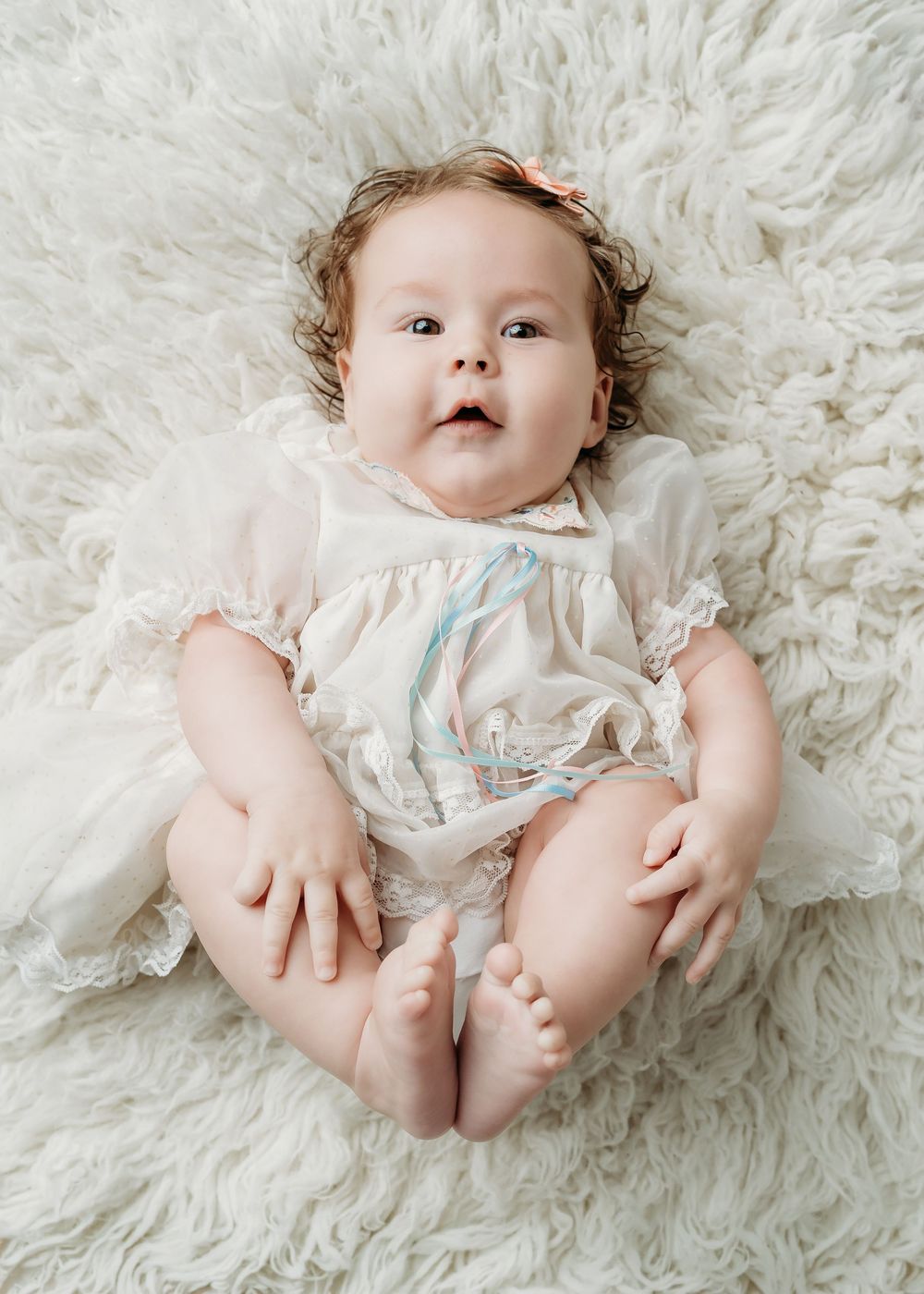Adorable baby girl with curly hair lying on a fluffy white rug wearing a delicate lace romper with a floral bow.