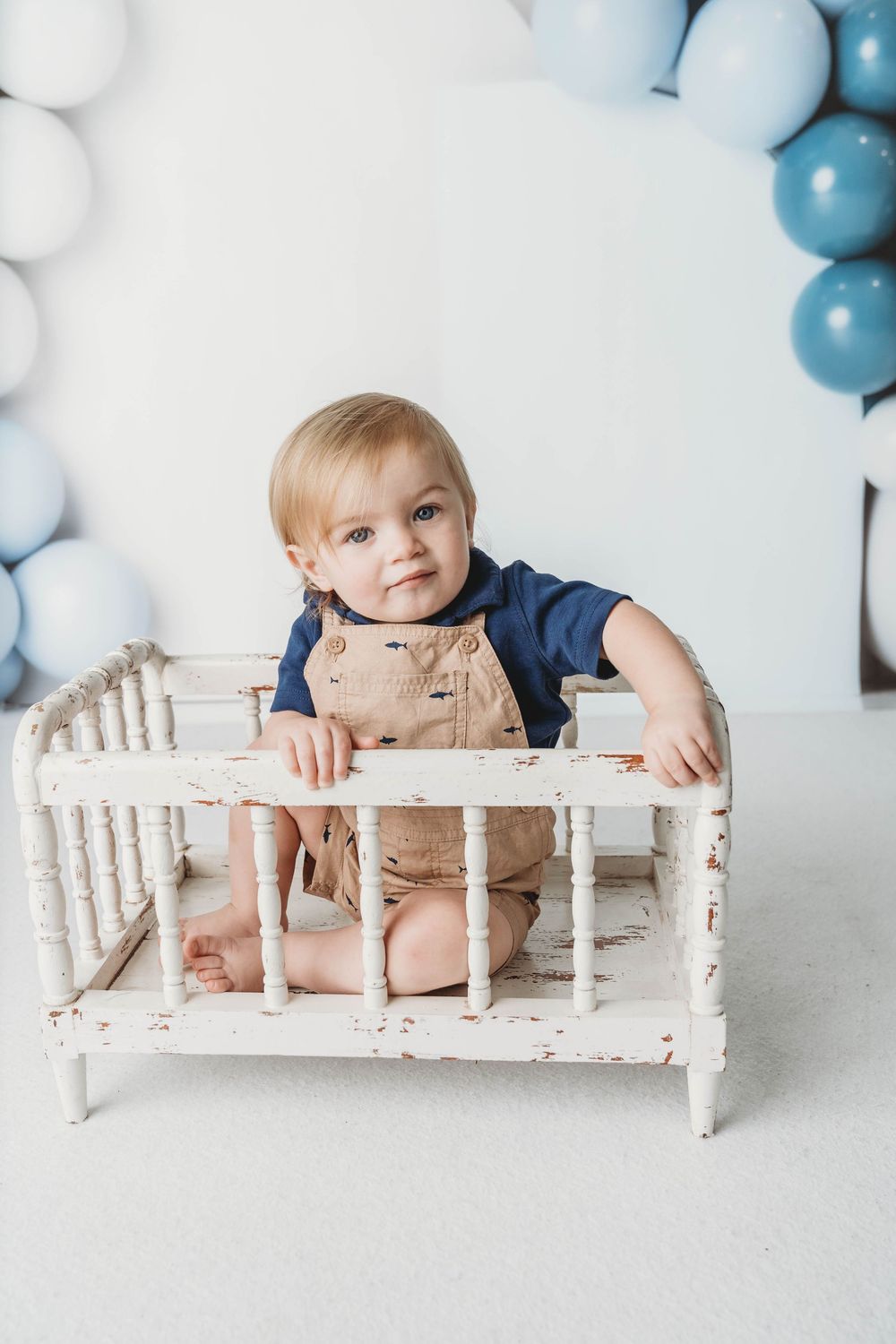 Cute toddler boy in tan overalls sitting in a vintage white mini crib surrounded by blue and white balloons.