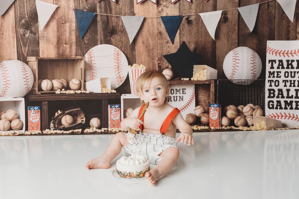 Baby boy in orange suspenders sits by smashed cake surrounded by baseball themed decorations and wooden backdrop.