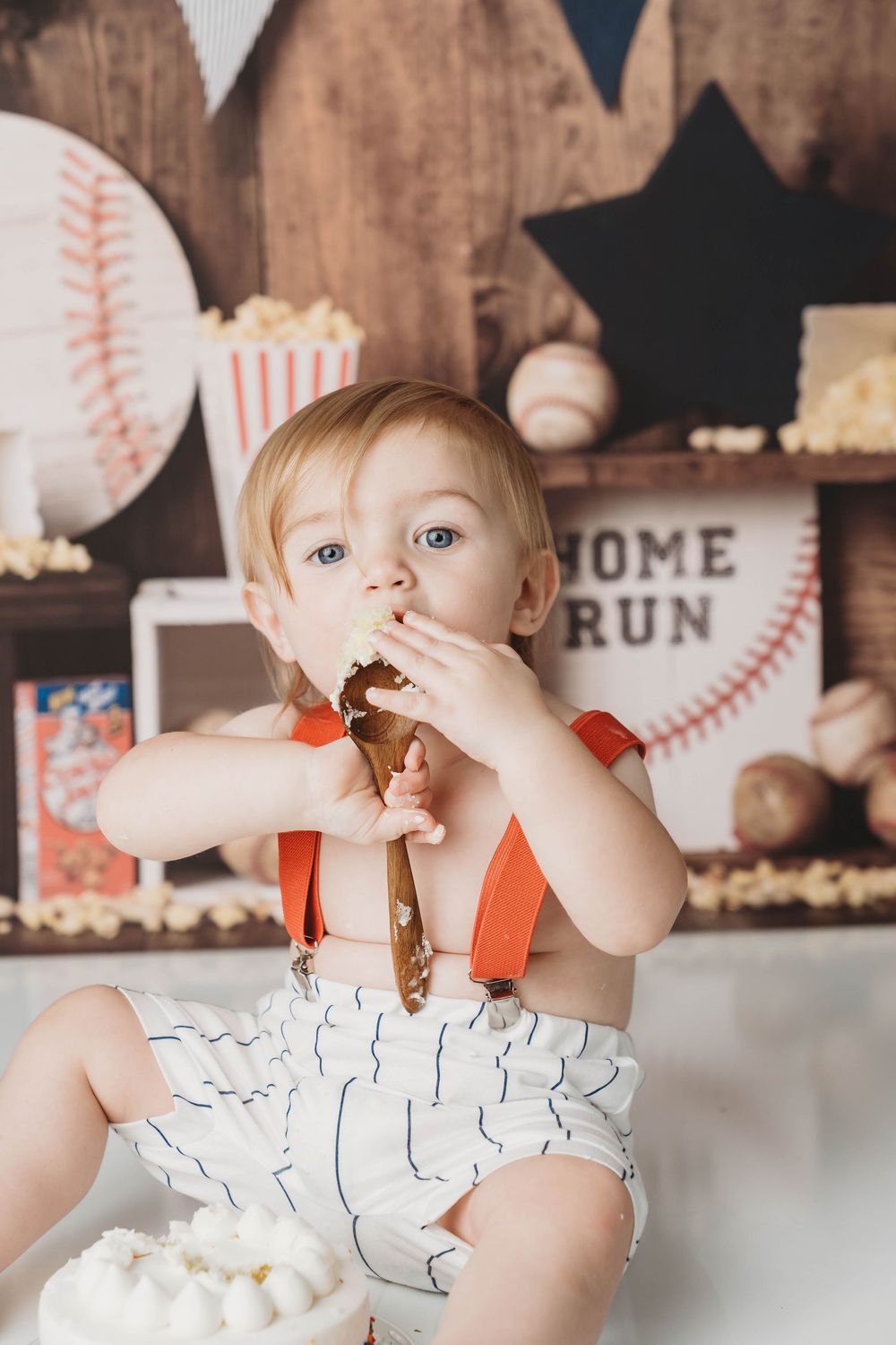 Baby boy in suspenders eating cake at a baseball-themed first birthday smash cake photoshoot.