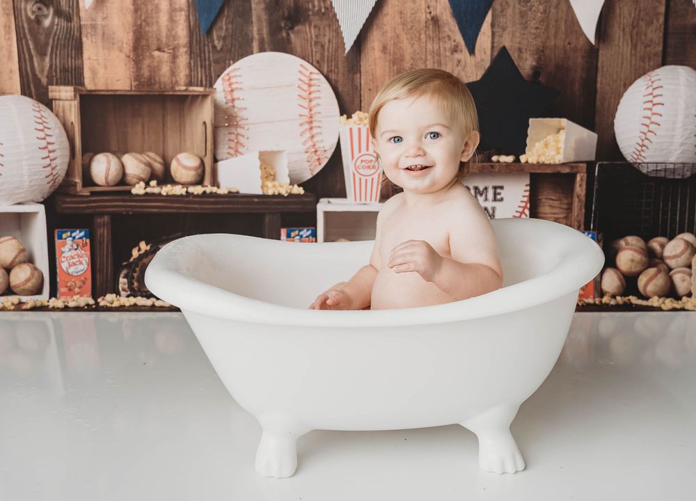 Smiling baby sitting in a white clawfoot tub surrounded by a baseball-themed backdrop with pennants and balls.