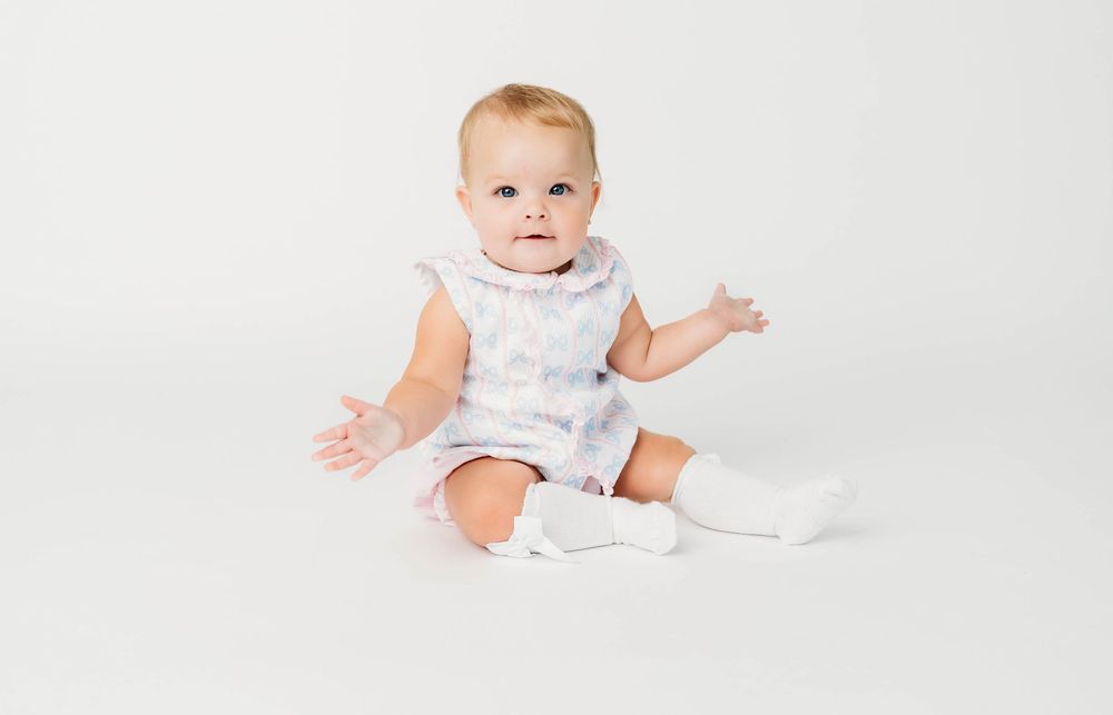 Baby sitting upright in white tie-dye romper and white socks on white background.