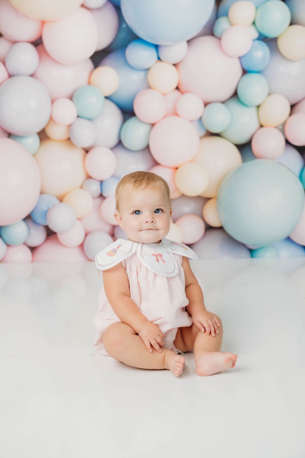 A baby girl in a pink monogrammed romper sits on white floor surrounded by pastel balloons backdrop.