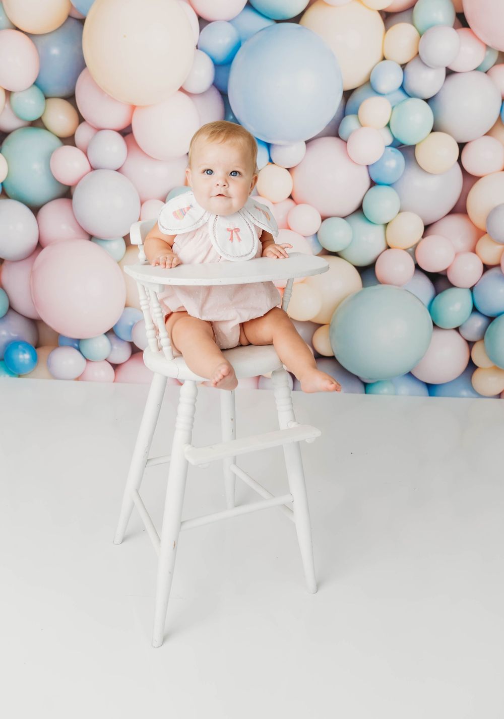 Baby in white high chair surrounded by pastel balloons backdrop, wearing pink outfit, first birthday photo session.