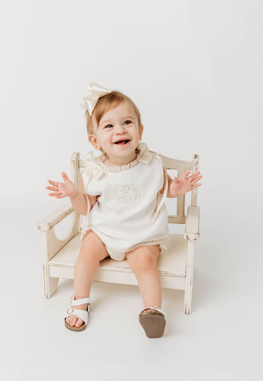 Smiling baby girl in cream monogram romper and bow headband sitting on white wooden chair.