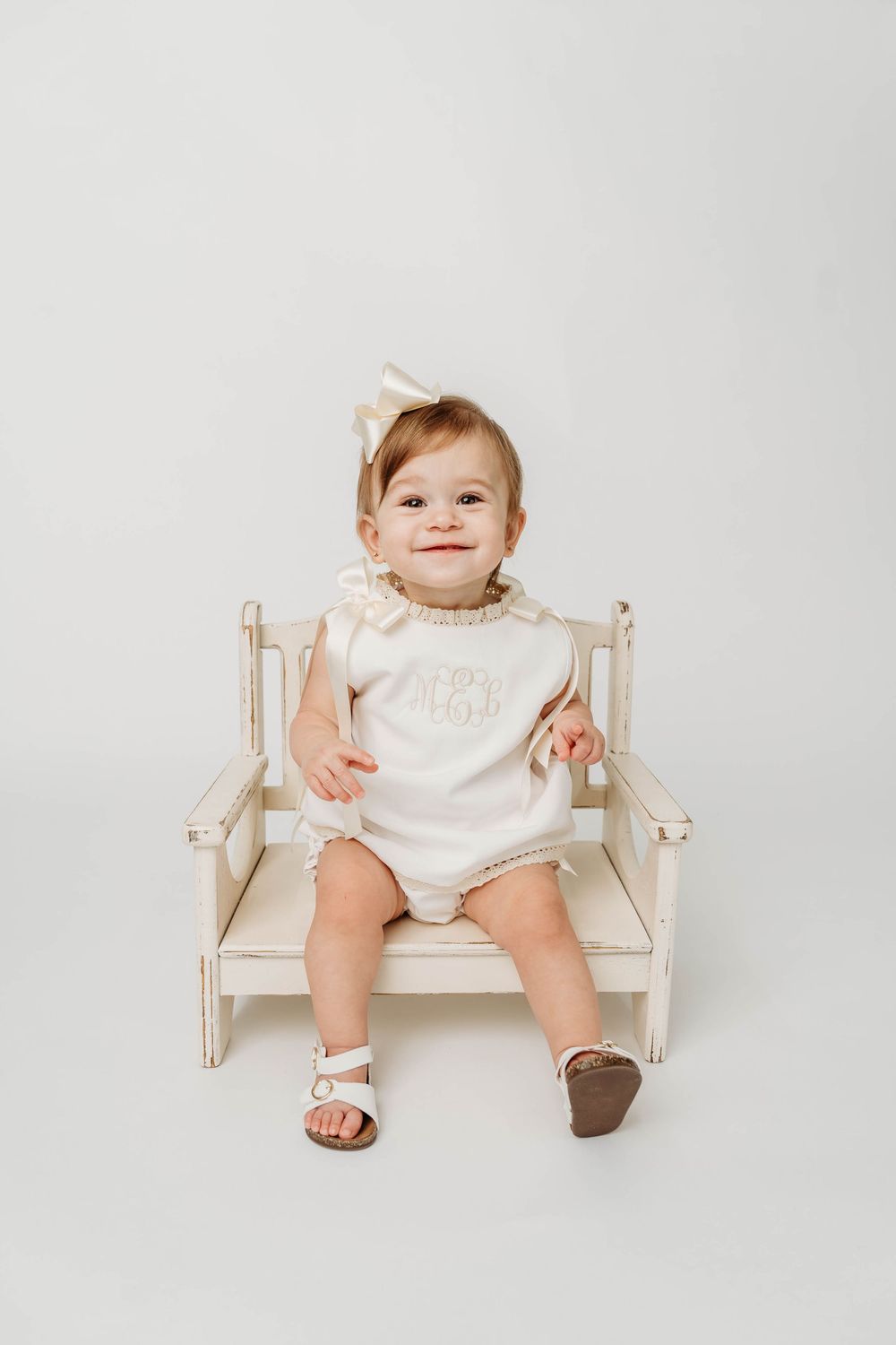 Smiling baby girl in cream monogrammed romper and bow headband sitting on a small wooden bench against white background.