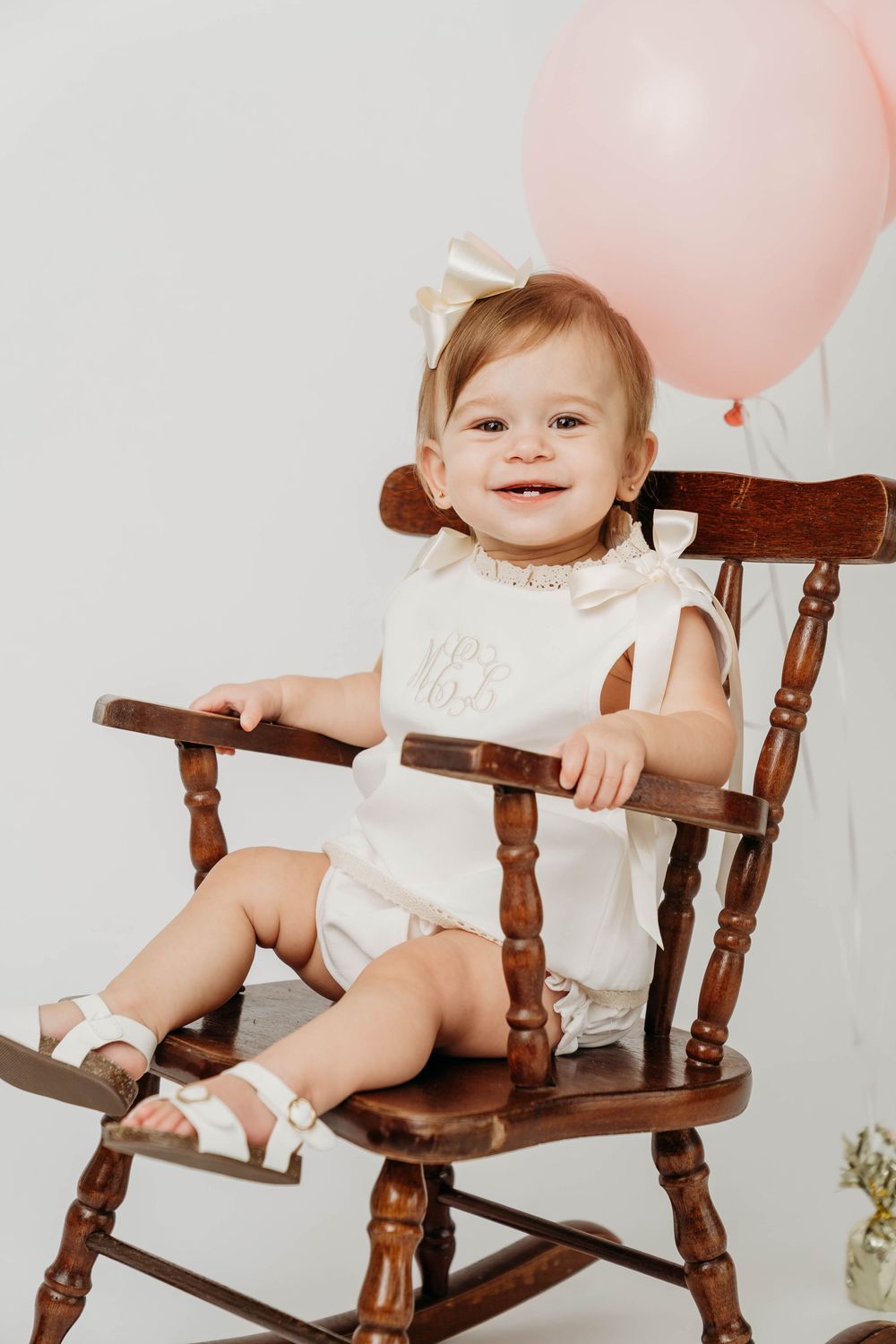 Smiling baby girl in white monogrammed romper sitting in wooden rocking chair with pink balloon.