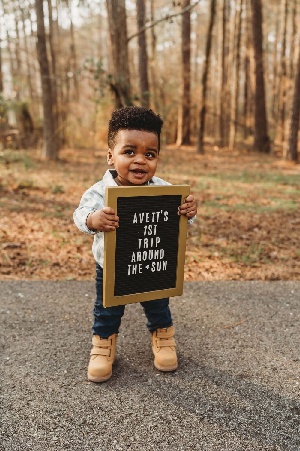 Smiling toddler boy holds a letter board sign reading Avett's 1st Trip Around the Sun outdoors.