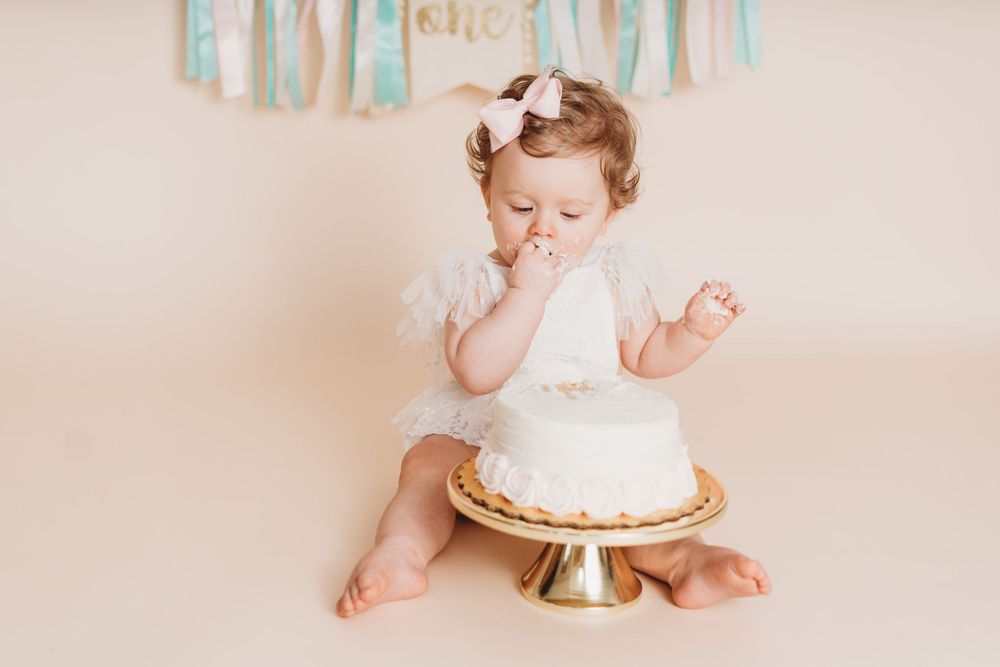Baby girl in white dress eating her first birthday cake smash on a gold cake stand against a cream background.