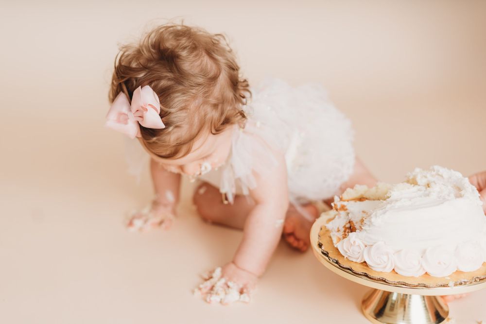 Baby girl in white tutu and pink bow smashes first birthday cake on beige studio backdrop.