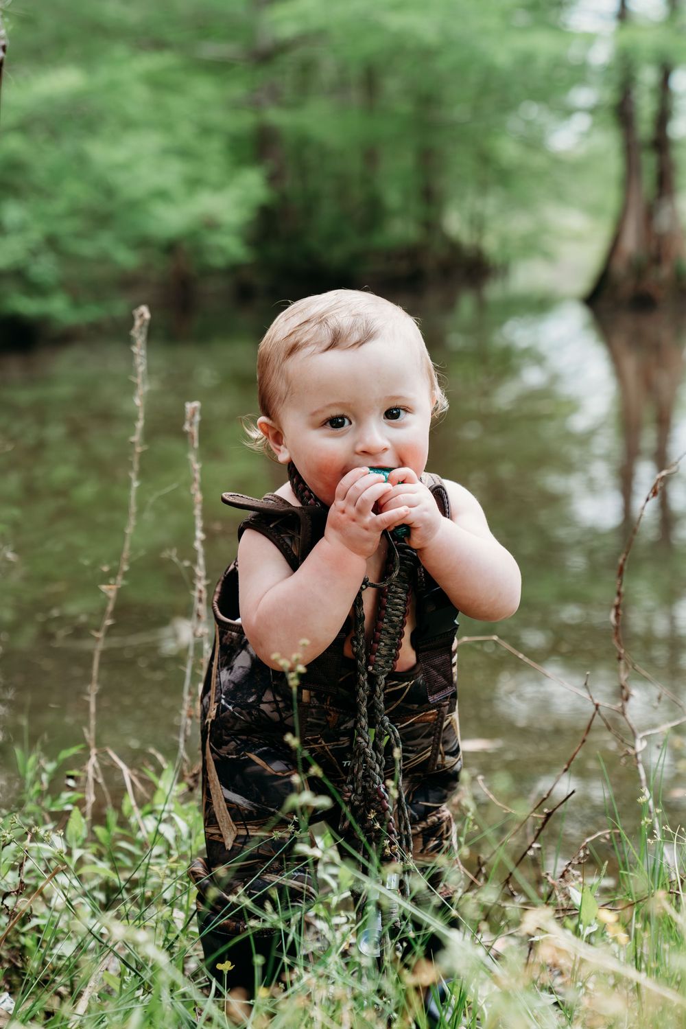 Toddler in camo outfit chewing on plants by a serene river surrounded by lush green vegetation.