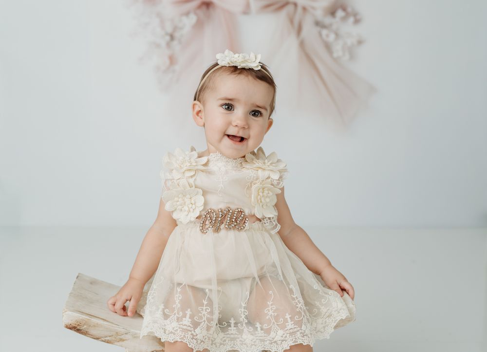Baby girl in ivory lace dress and floral headband smiling during professional photo session.
