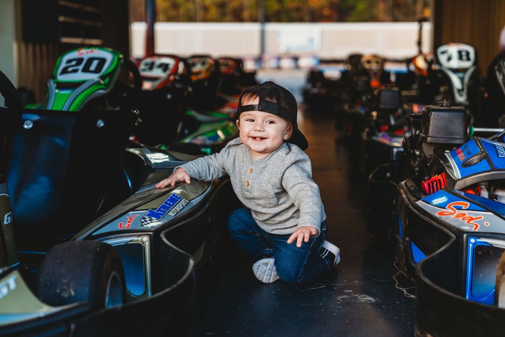 Smiling toddler in a baseball cap sitting between go-karts at an indoor karting track.