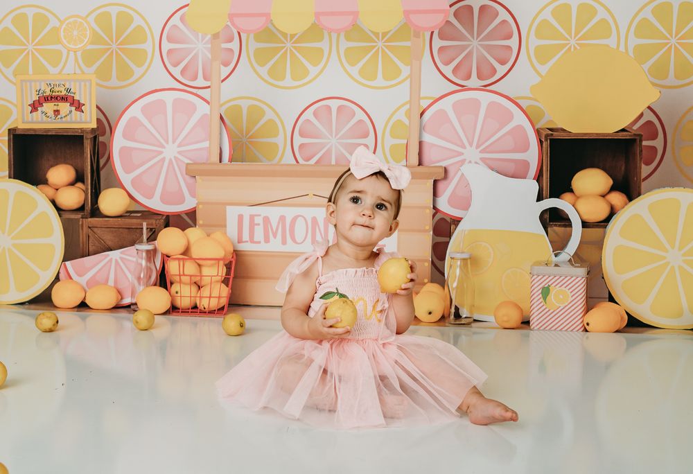 Baby girl in pink tutu sits among lemons with lemonade stand backdrop for a fun first birthday cake smash photo.