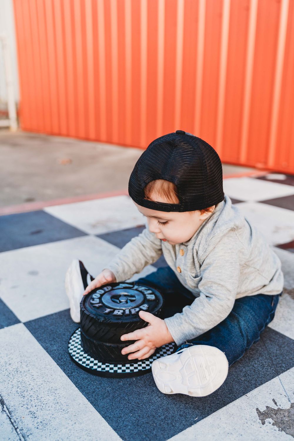 Toddler boy in black cap and jeans sitting on checkered floor playing with a skateboard wheel outdoors.