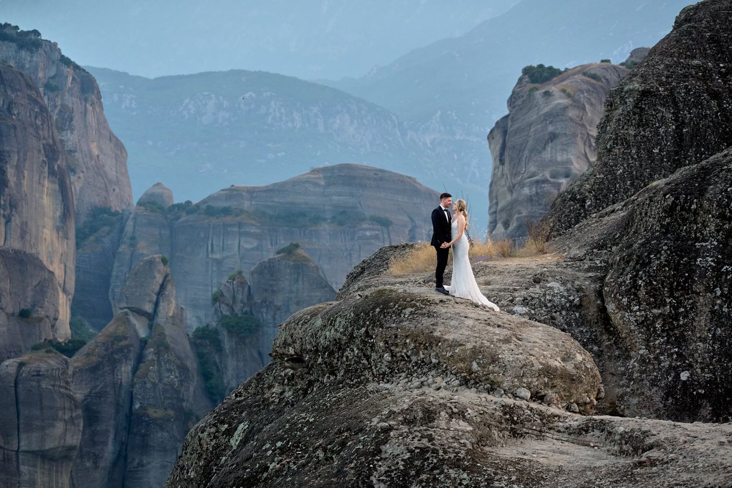 Wedding couple embracing on a rocky cliff in Meteora, Greece, with mountains in background.