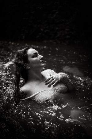 Black and white photo of a woman emerging from dark water, looking upward with wet hair and hands crossed over chest.