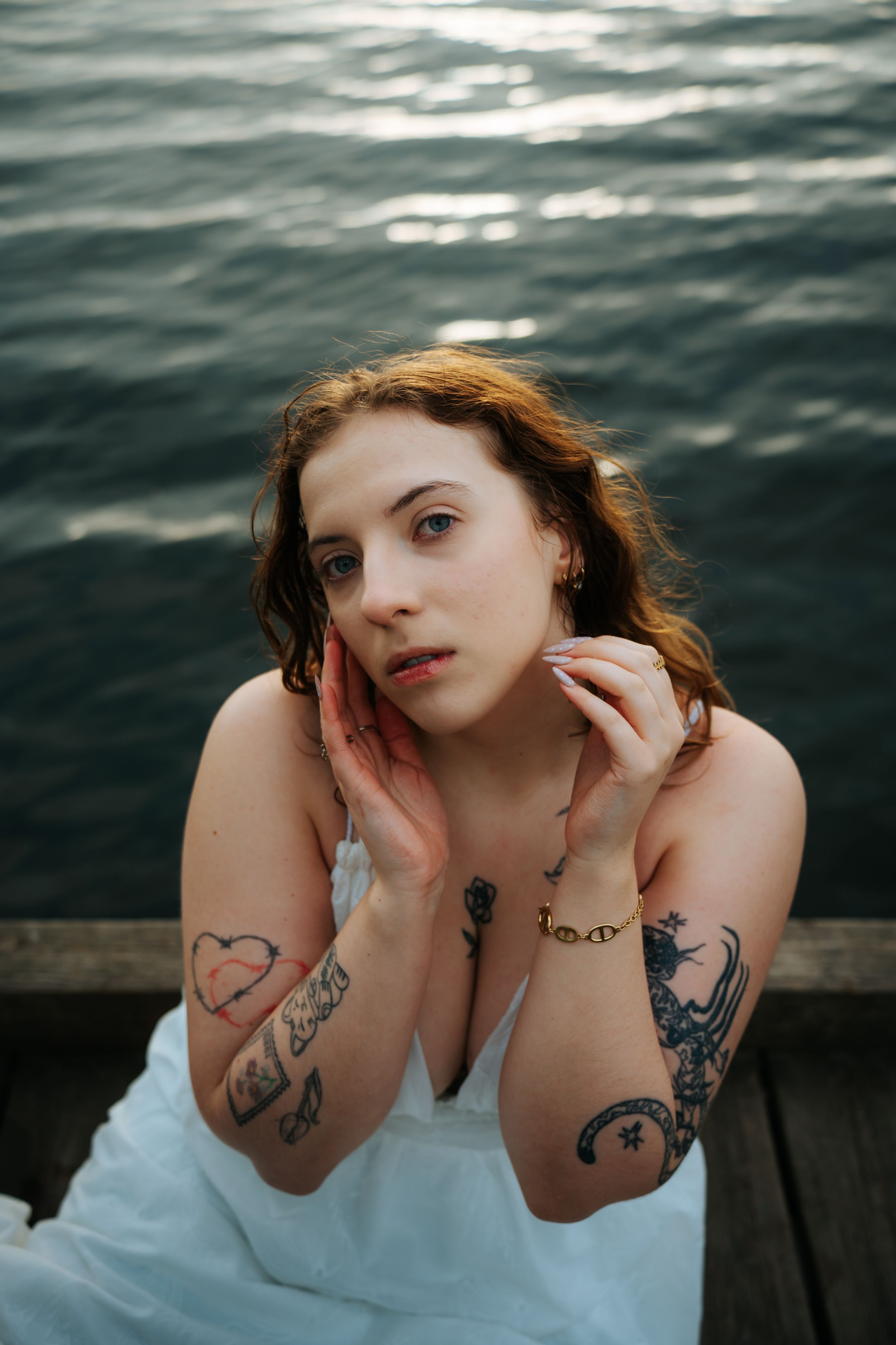 Young tattooed woman in white dress poses by water on dock, hands framing her face, with visible arm tattoos.