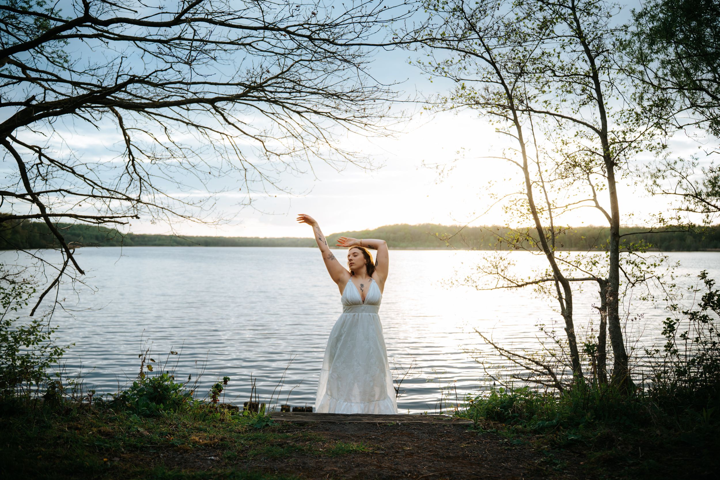Woman in white dress dancing by a sunlit lake at sunset, surrounded by trees in a serene outdoor setting.