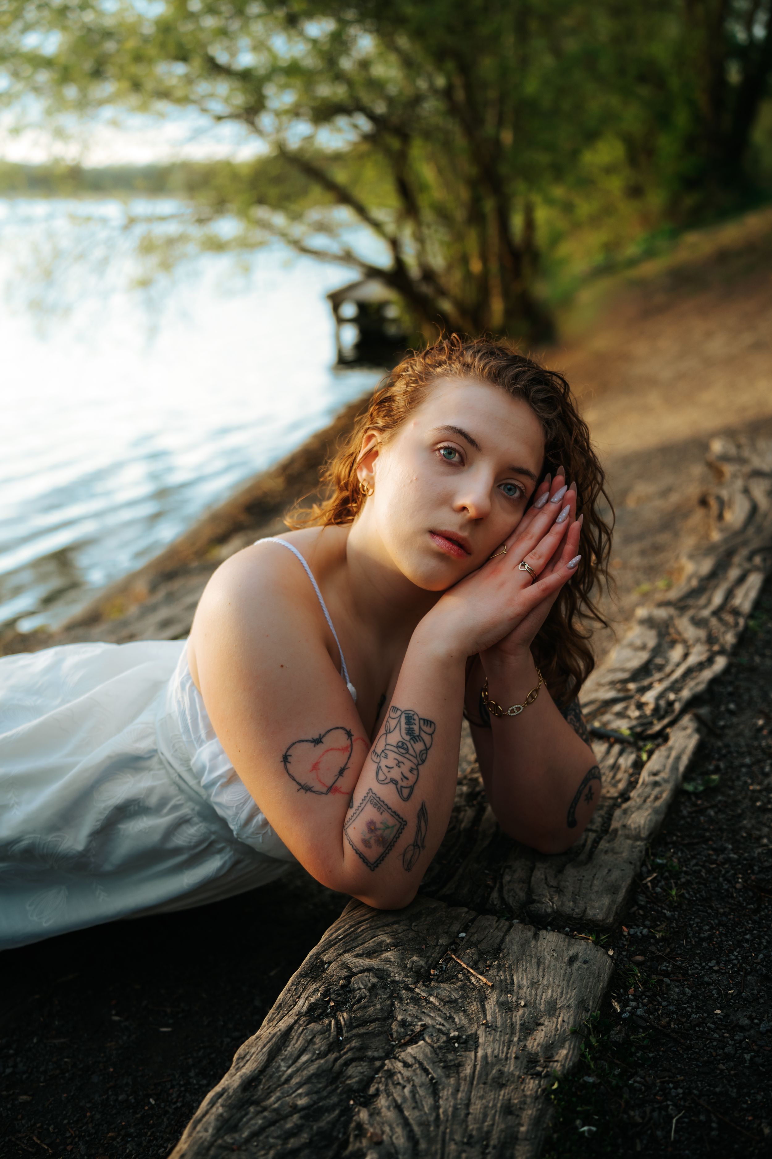 Young woman with tattoos in white dress resting on a log by a serene lakeside at golden hour.