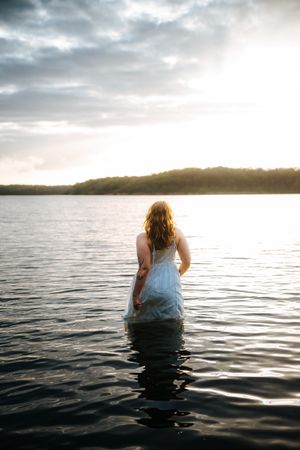 Woman in white dress wading in calm lake at golden sunset with cloudy sky and tree-lined shore.