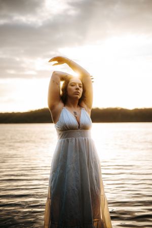 Woman in white dress standing in water at golden sunset, arms raised gracefully above head with eyes closed.
