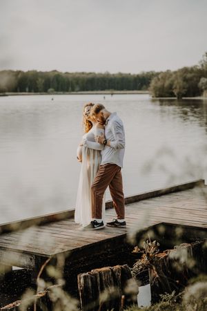 Couple sharing a tender kiss on a wooden dock by a serene lake, woman in white dress with visible baby bump.