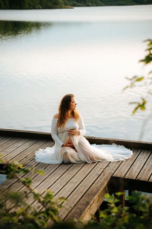 Pregnant woman in white gown sitting on wooden dock by serene lake, cradling baby bump at sunset.