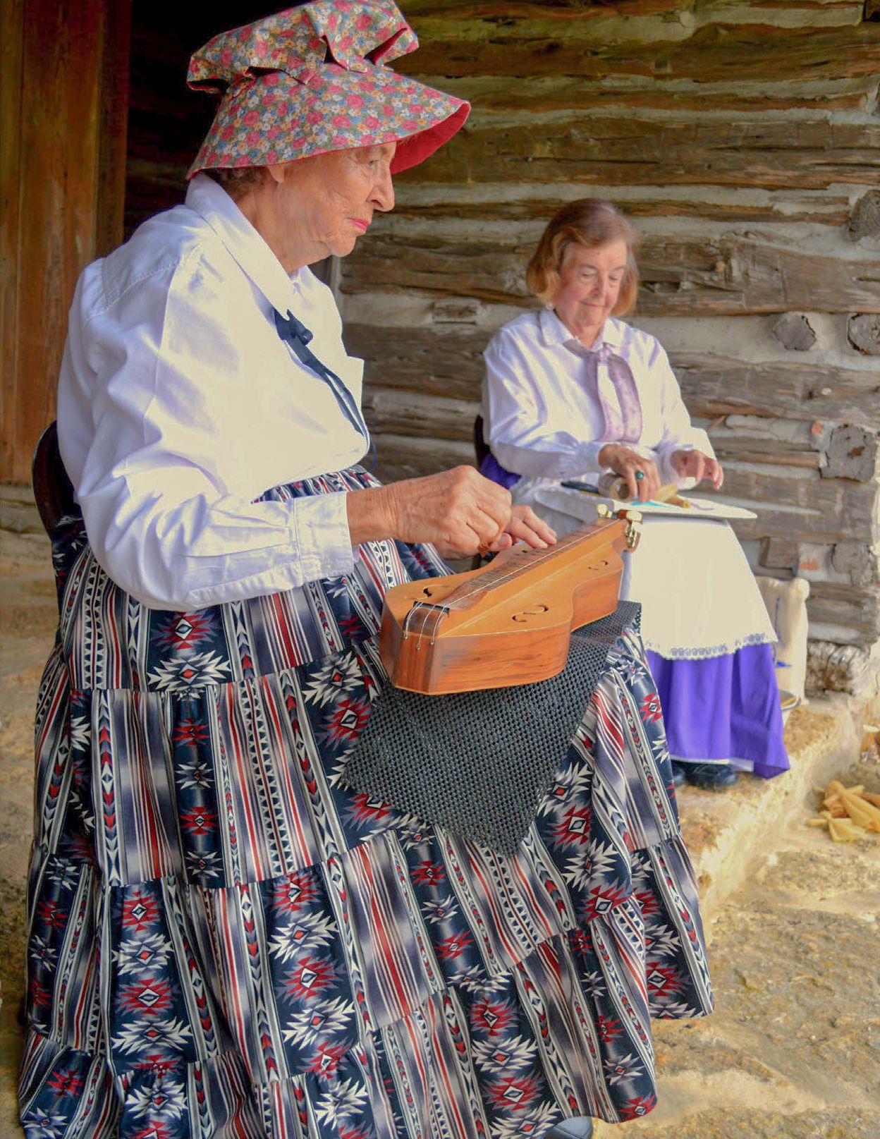 Two elderly women play instruments on a porch while dressed in historic dresses