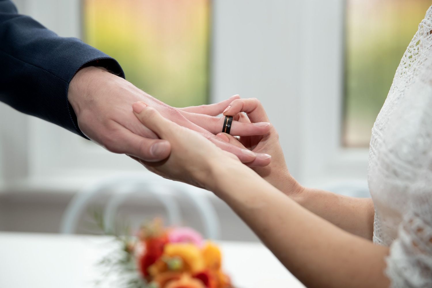 Groom placing a wedding ring on bride's finger during ceremony, with colorful flowers in the background.