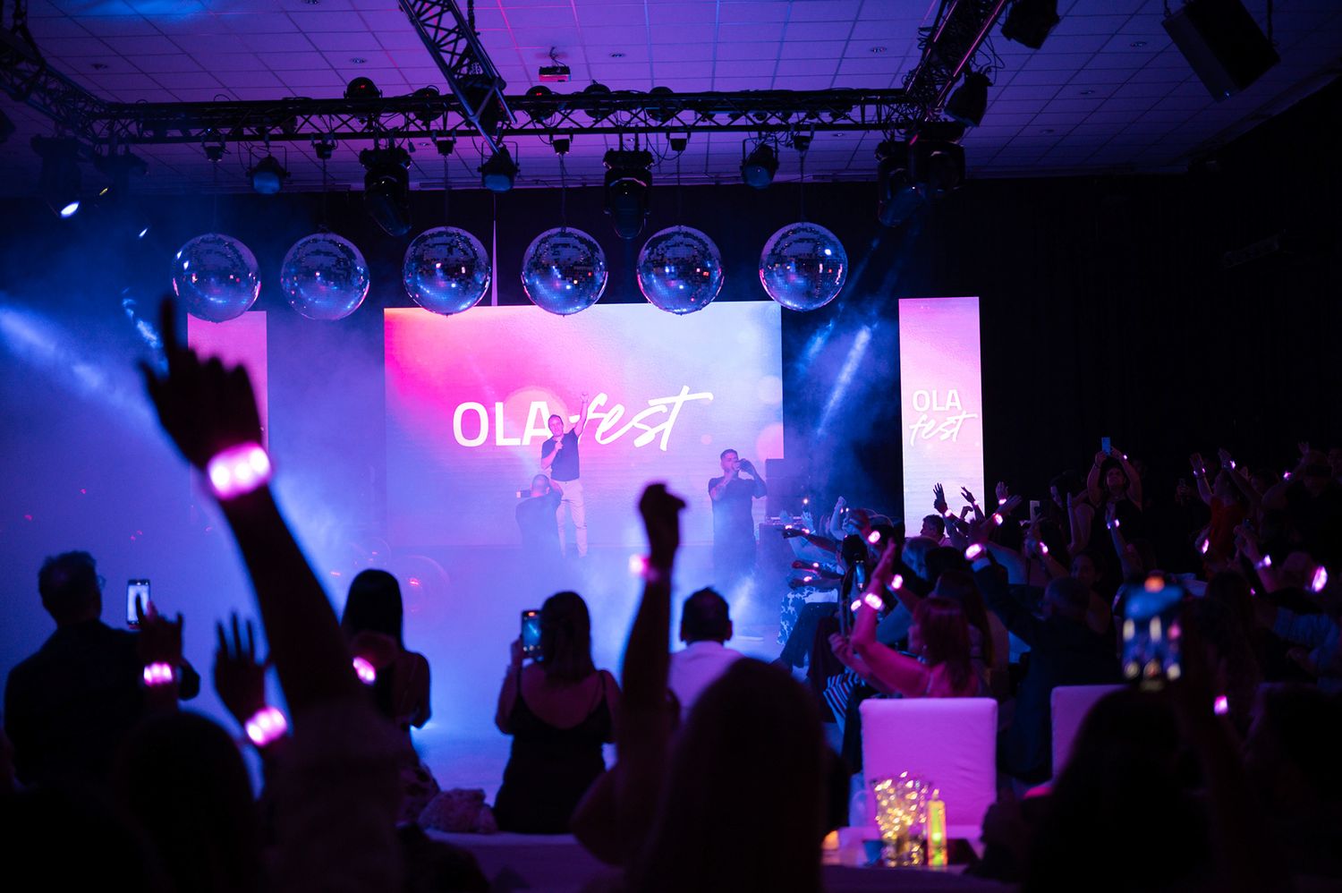 Energetic concert with crowd holding glowing wristbands under disco balls and stage lights at an OLA event.