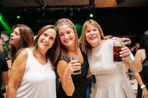 Three women smiling and holding drinks at a lively nightclub event with green stage lighting.