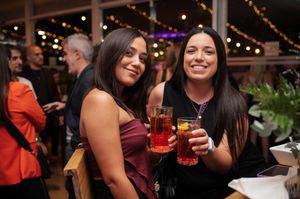 Two women smiling and holding red cocktails at an outdoor evening social event.